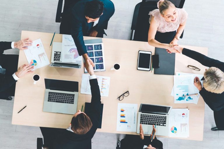 Mehrere Personen in Anzügen sitzen um einen Tisch und diskutieren, während sie sich die Hände schütteln.